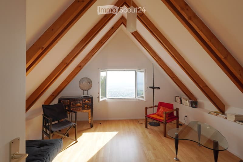 Bright attic room with wooden beams, large window offering a view, sofa, chairs, glass coffee table, floor lamp, and decorative cabinet.