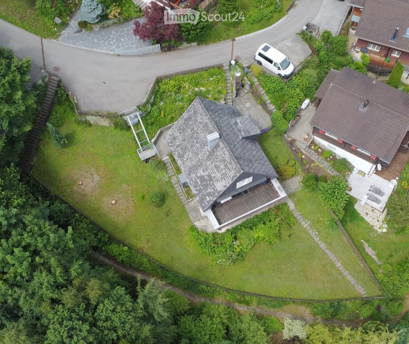 Aerial view of a two-story house with a grey roof, surrounded by a well-maintained garden and driveway. The house features a wooden deck and patio area, with neighboring houses and a parked van visible in the background.