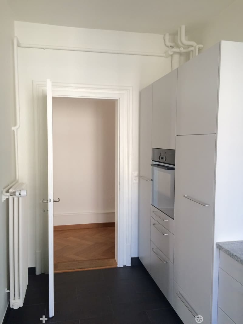 A narrow hallway with white walls, black tiled floor, and a radiator on the left side. An open doorway leads to another room with wooden flooring.