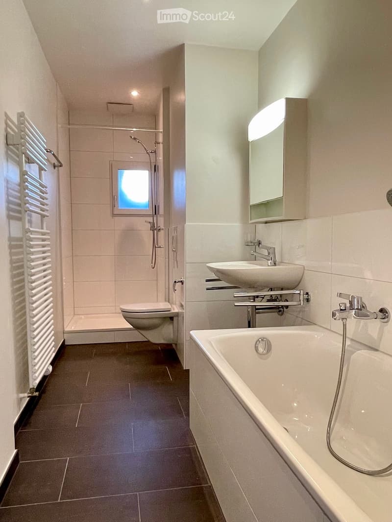 Modern bathroom with a bathtub, sink, toilet, and a radiator. Features a large mirror above the sink and a window in the shower area.