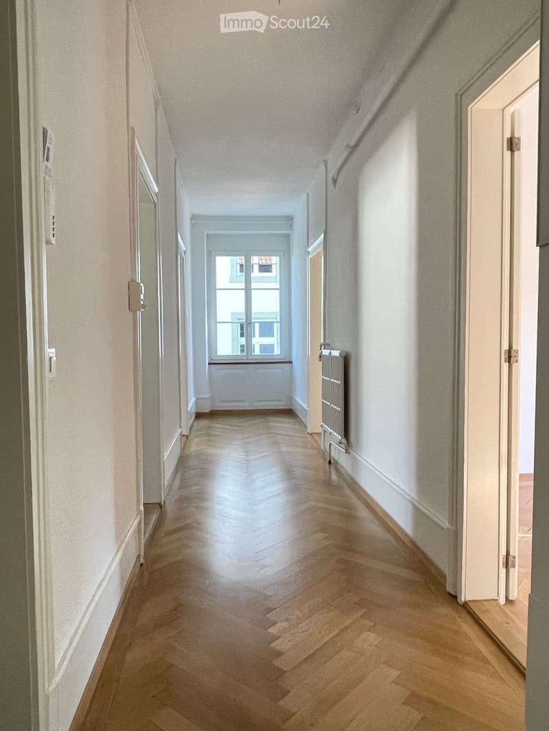 A long, narrow hallway with wooden flooring and white walls, featuring a radiator on the right side and a window at the far end.
