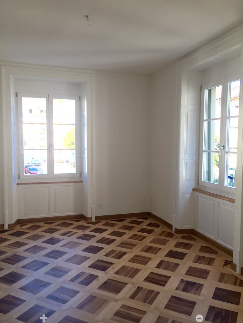 Empty room with wooden floor and large windows, featuring white walls and radiators.