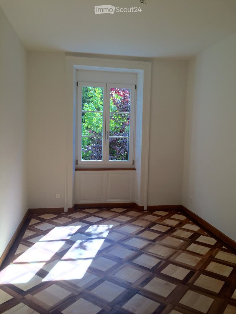 Empty room with light-colored walls, wooden floor, and a large window with double doors leading outside.
