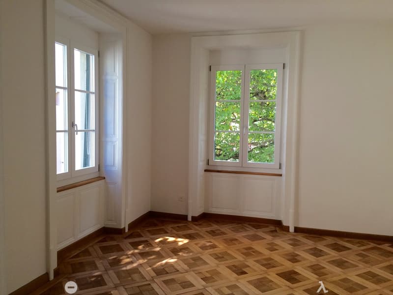 Empty room with wooden parquet flooring, white walls, and large windows with white frames. The room has a door and radiator covers, with natural light streaming in.