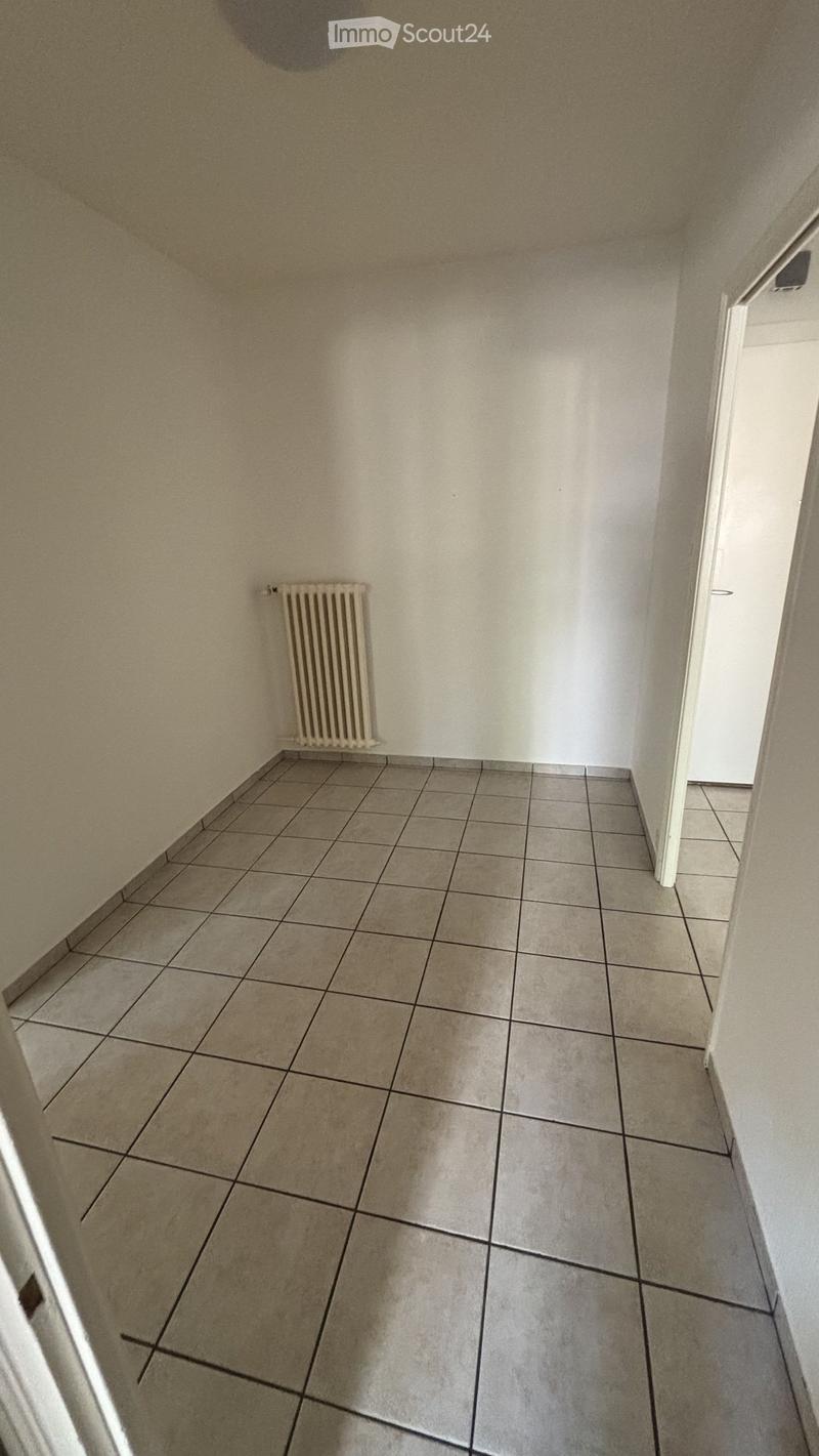 Empty hallway with tiled floor and white walls, featuring a radiator on one side and a partially open door at the end.