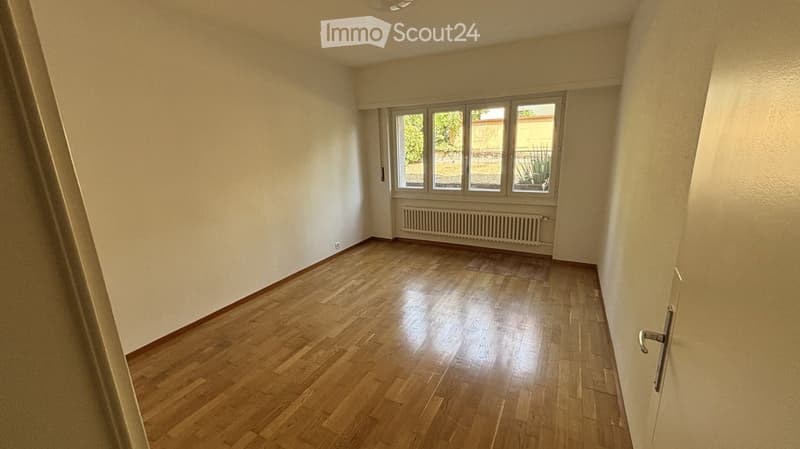 Empty room with wooden flooring, white walls, and large windows offering a view of greenery outside. A radiator is visible below the windows.