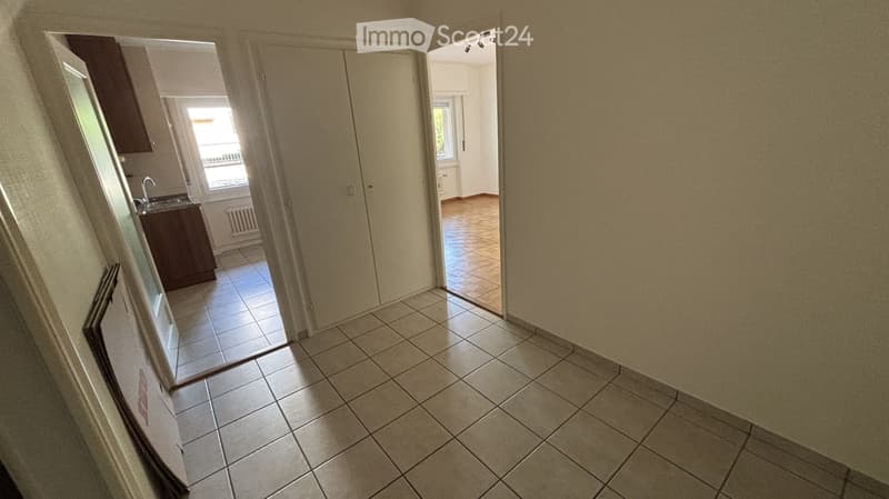 Empty hallway with tiled flooring, leading to a kitchen and another room. The walls are light-colored, and the space is well-lit with natural light coming from the adjacent rooms.