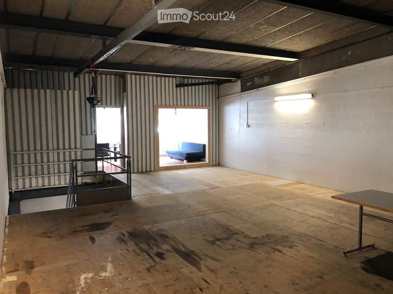 An empty, industrial-style room with exposed metal beams, a metal staircase, and a large open doorway leading to another room with a sofa. The floor is unfinished concrete, and the walls are white with some visible wear.