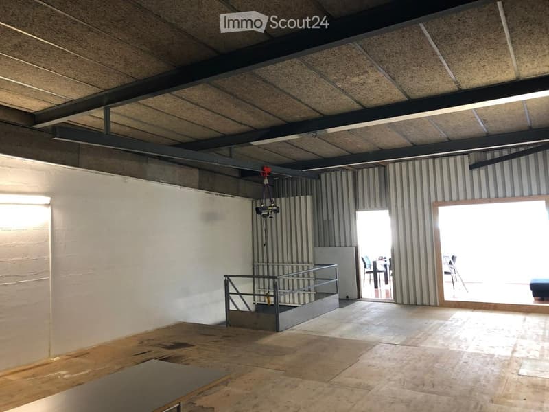 Empty room with exposed metal beams, white walls, and a staircase leading to another area. The floor is tiled, and there is a large open doorway leading to an outdoor area.