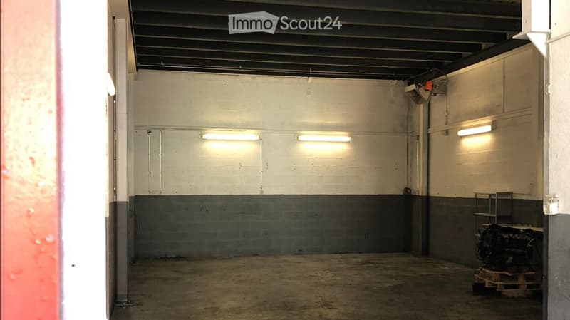 Empty garage with concrete floor, white walls, fluorescent lighting, and a small storage area with a workbench and tools.