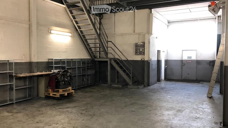 Empty industrial-style room with concrete floor, metal shelving, metal staircase, and a small work area with a table and engine on a pallet.