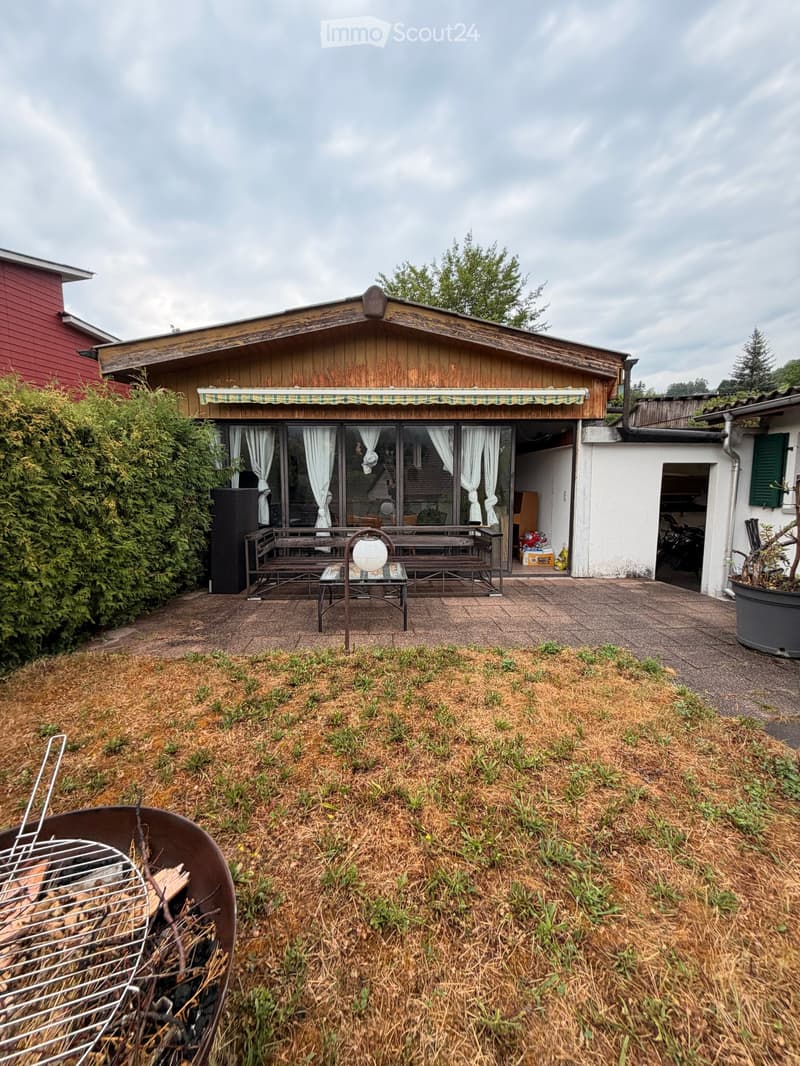 Outdoor patio area with wooden bench, table, grill, and a covered wooden structure with large windows and curtains.