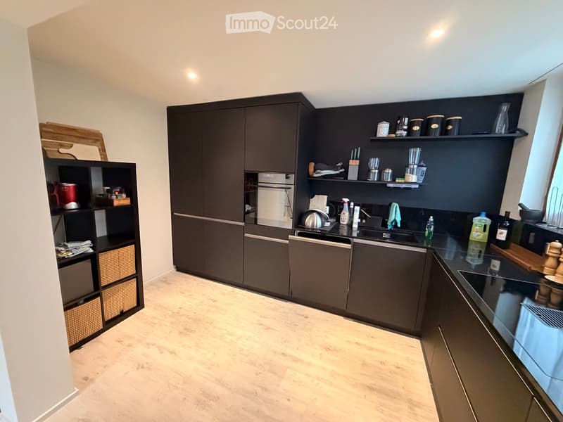 Modern kitchen with dark wooden cabinets, stainless steel appliances, and a black countertop. Includes shelves with various kitchen items, a wicker basket unit, and a wooden cutting board on display.