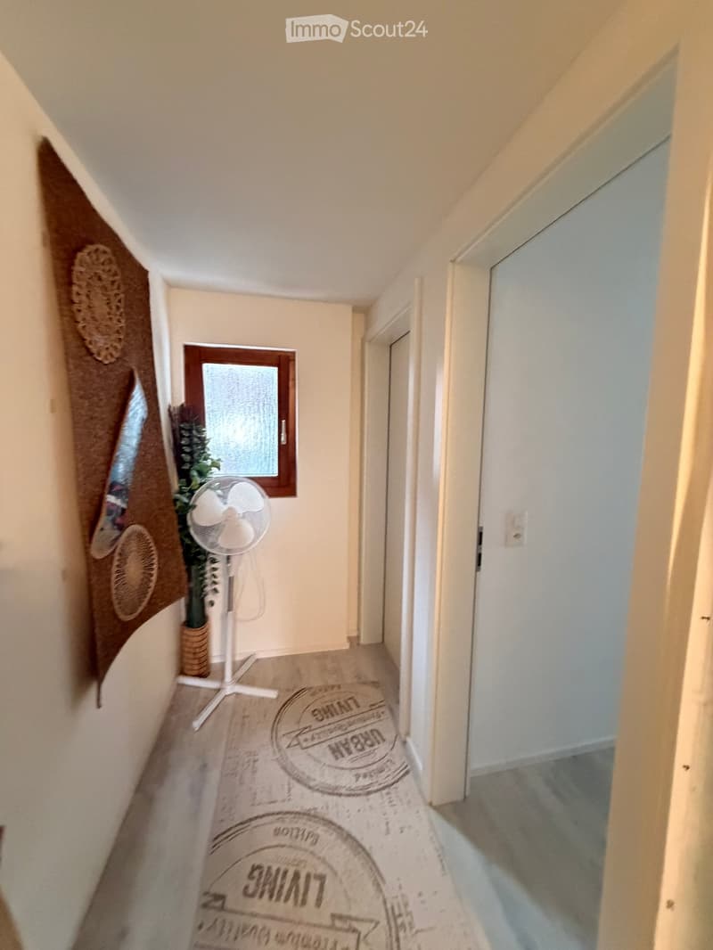 A narrow hallway with light-colored walls, carpeted floor, and a small window. Features include a fan, a potted plant, and a decorative wall hanging on the left side.