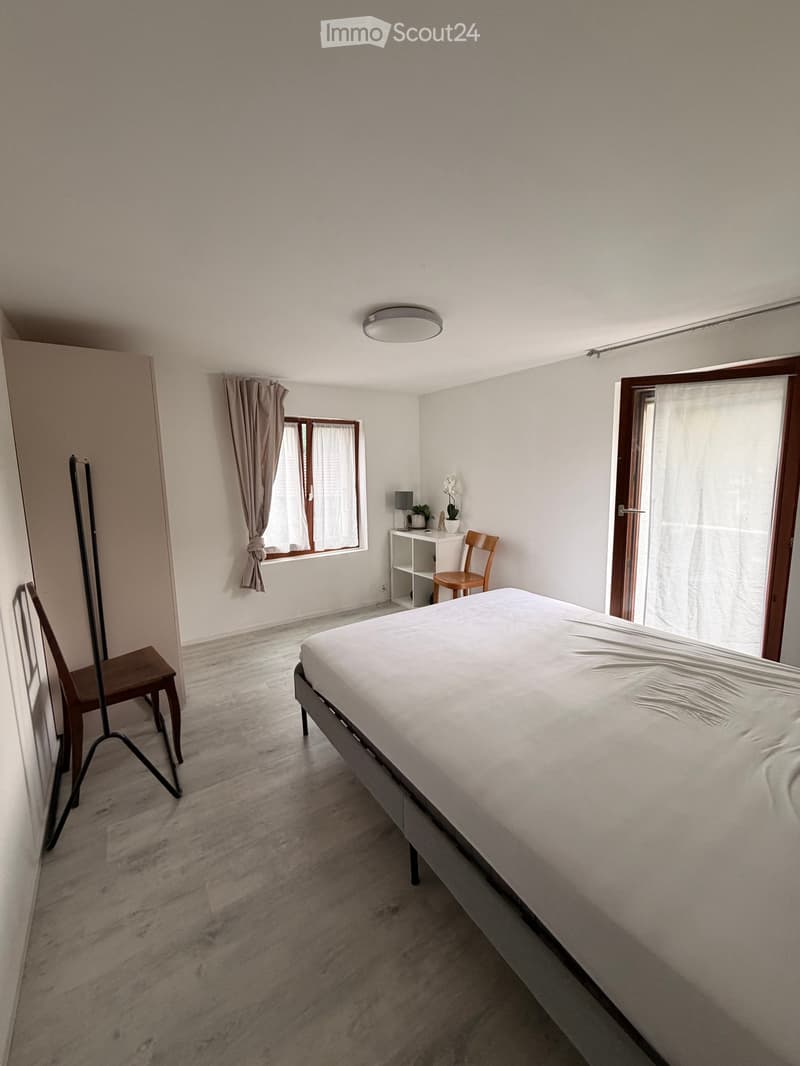 Minimalist bedroom with a single bed, two wooden chairs, a small table with a lamp and plant, and a window with curtains.