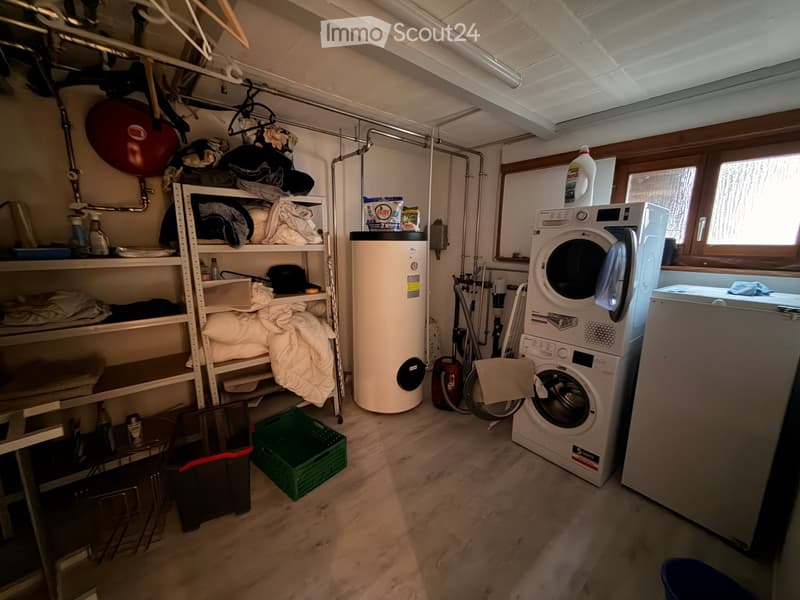 Laundry room with a washing machine, water heater, shelves filled with linens and cleaning supplies, and a green laundry basket on the floor.