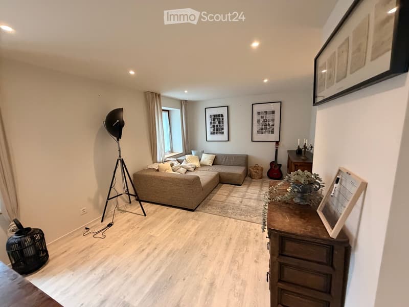 Modern living room with light wooden flooring, a grey sectional sofa, a floor lamp, a guitar, a small table with a vase, and framed artwork on the walls.