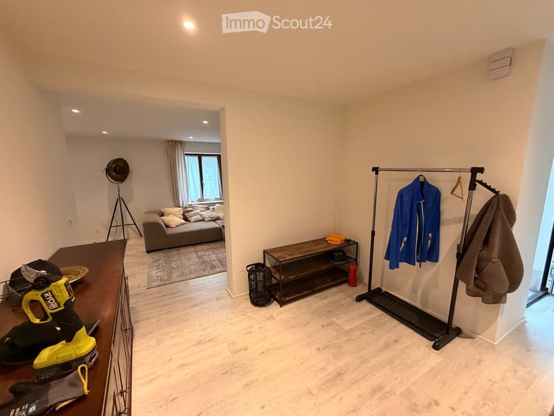 A modern, well-lit living space with a sofa, a wooden table, a floor lamp, a coat rack, and various items including a power tool and a fire extinguisher.