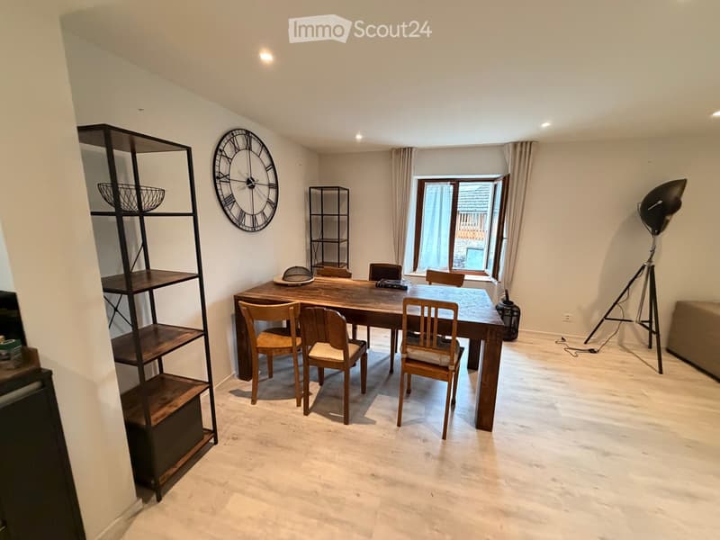 Dining room with a wooden dining table, six chairs, a large wall clock, a tall black shelving unit, and a floor lamp near the window.