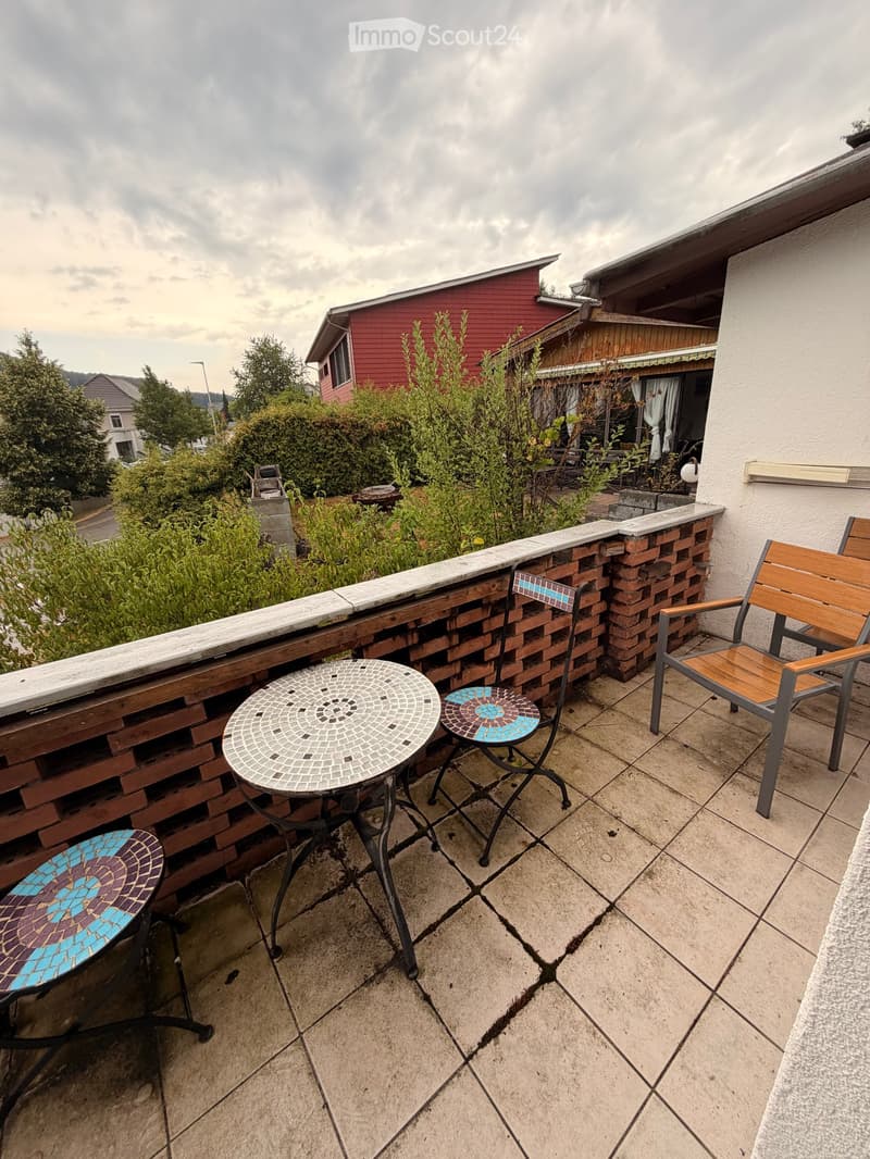 Balcony with tiled tables, chairs, and a brick privacy wall, overlooking a residential area with greenery.