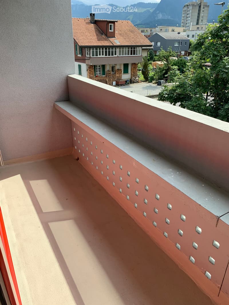 Balcony with a red railing and decorative cutouts, overlooking a residential area with houses, trees, and mountains in the background.