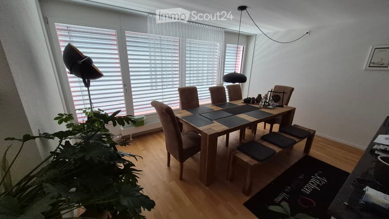 Bright dining area with a wooden dining table, six chairs, a bench, and a hanging lamp. Large windows with blinds and a potted plant are visible.