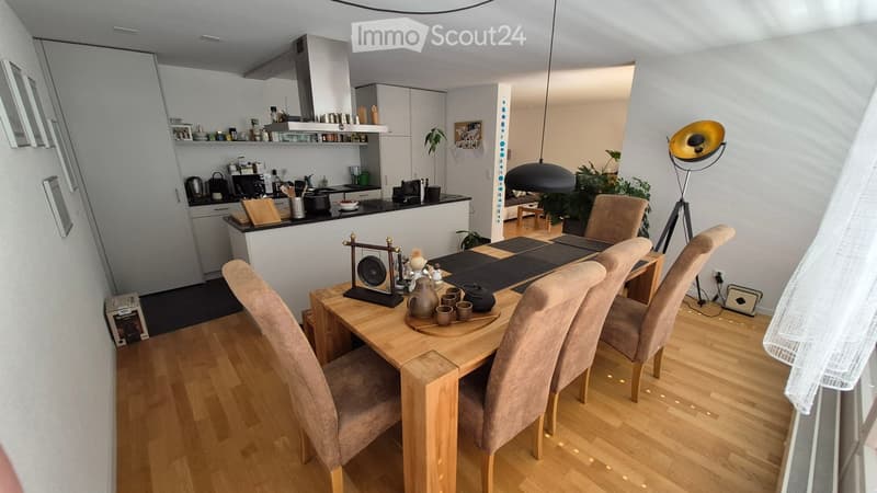 Open-plan living and dining area with a wooden dining table surrounded by six chairs, a modern kitchen with white cabinets and stainless steel appliances, pendant lighting, and a floor lamp.