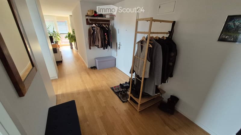A well-lit hallway with wooden flooring, coat rack, shoe rack, and storage bench. The hallway leads to a living area with plants and a sofa visible in the background.
