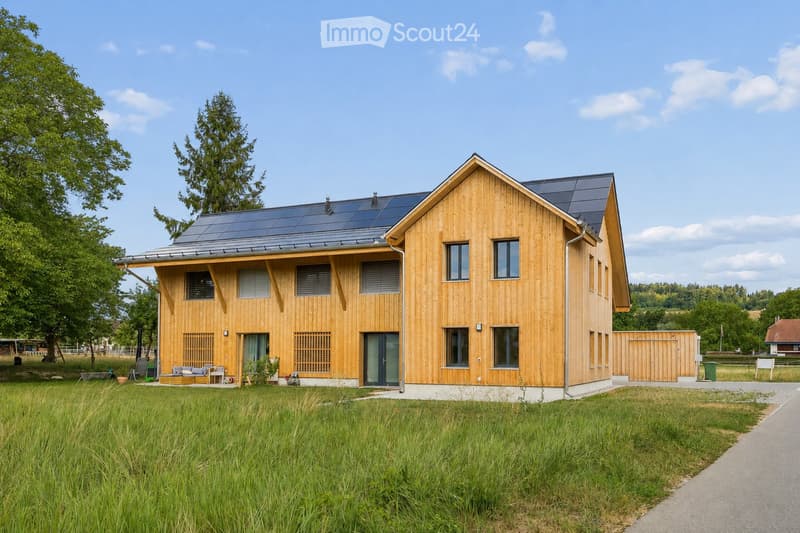 Modern two-story wooden house with solar panels on the roof, large windows, and a spacious front yard with a small seating area.