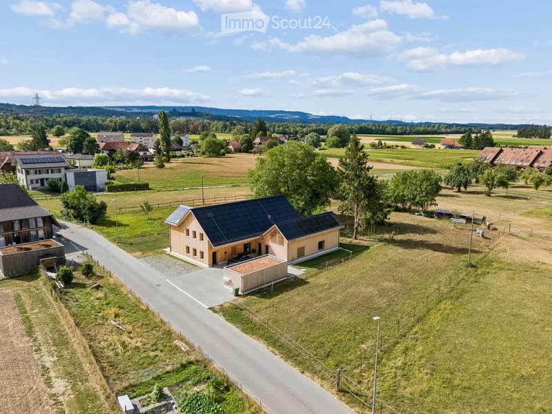 Aerial view of a modern, two-story house with a dark roof and solar panels, surrounded by a rural landscape with fields, trees, and neighboring houses.
