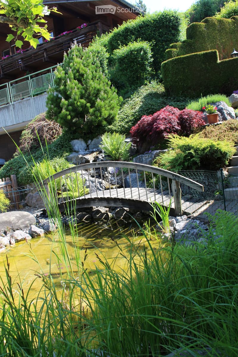 Garden with a pond, wooden bridge, various plants, shrubs, and a small waterfall.