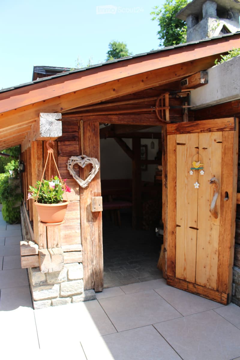 Wooden shed with a tiled roof, stone base, and decorative elements including a hanging flower pot and a heart-shaped wooden decoration.