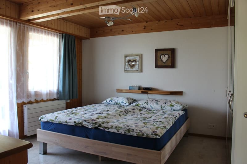 Cozy bedroom with wooden bed, floral bedding, wooden shelf, framed pictures, and sheer curtains on the window.