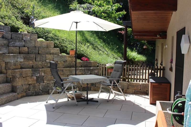 Patio area with a stone wall, tiled floor, outdoor table and chairs, umbrella, and greenery in the background.