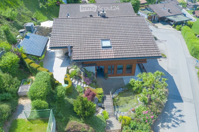 A two-story house with a tiled roof, surrounded by a well-maintained garden, a small pond, and a driveway leading to a garage. The house features large windows and a wooden facade on the lower level.