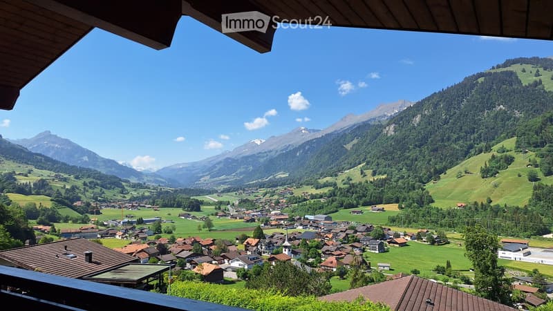 Panoramic view of a village nestled in a valley surrounded by lush green hills and snow-capped mountains under a clear blue sky.