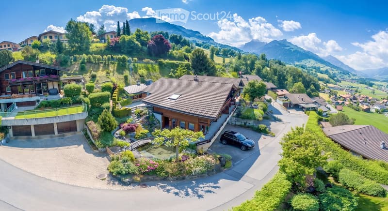 Aerial view of a large, modern house with a garage, surrounded by lush greenery and a scenic mountain backdrop.