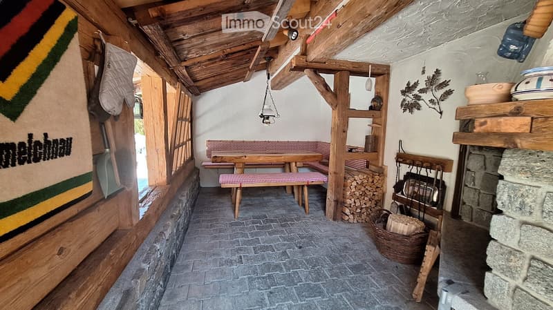 The image shows a rustic wooden room with a stone fireplace, wooden beams, and a wooden table with benches. There is a wood stack, a basket, and a wooden shelf with various items. The room has a cozy, traditional design with wooden walls and a wooden ceil