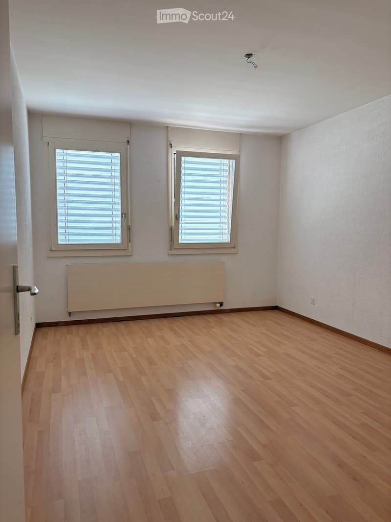 Empty room with wooden flooring, white walls, two windows with blinds, a radiator, and a door.
