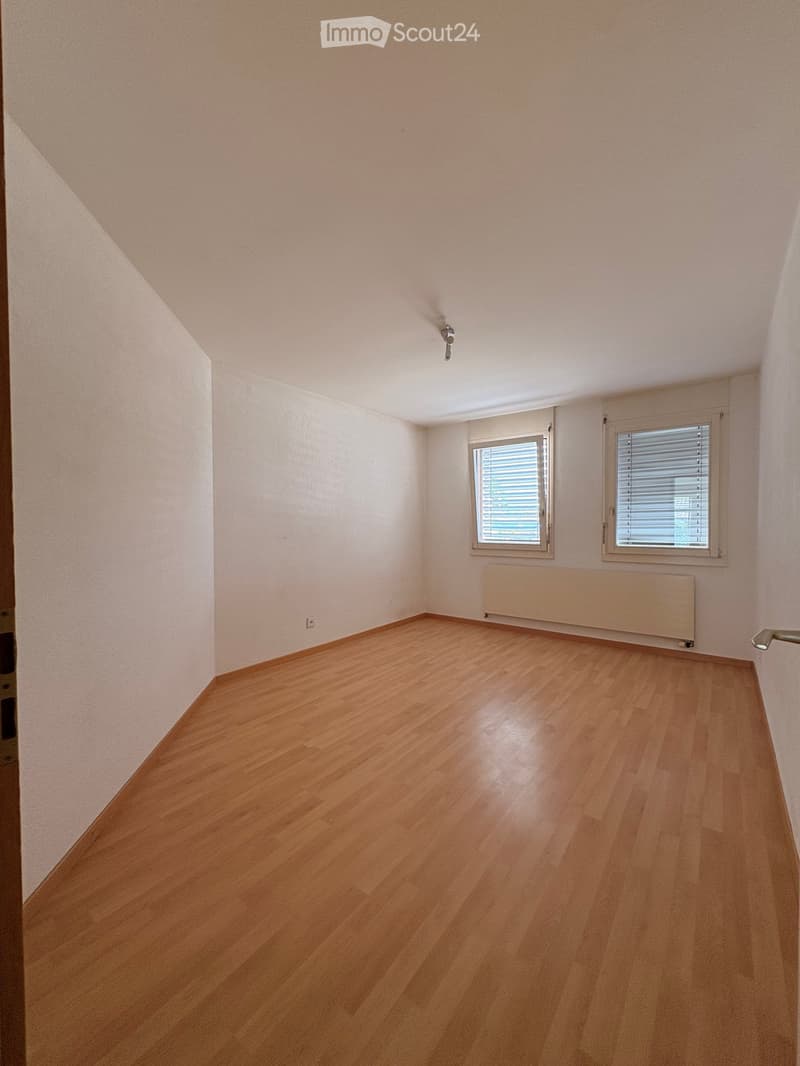 Empty room with wooden flooring, white walls, two windows with blinds, and a radiator under one window.