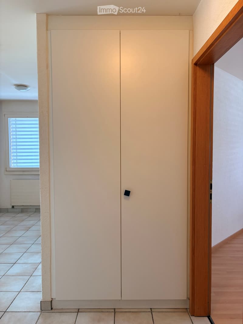 Empty hallway with tiled floor, white walls, and a closed white cabinet. A wooden door frame leads to another room with wooden flooring visible.