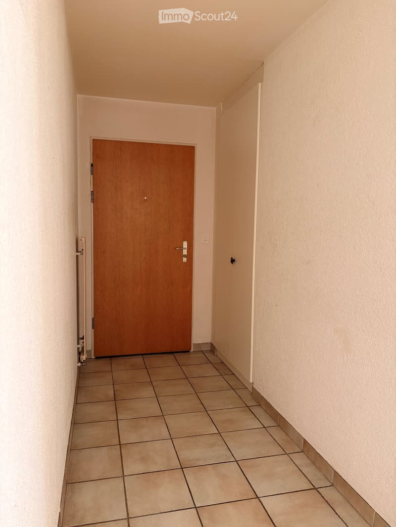 Empty hallway with tiled floor, beige walls, and a wooden door on the left side.