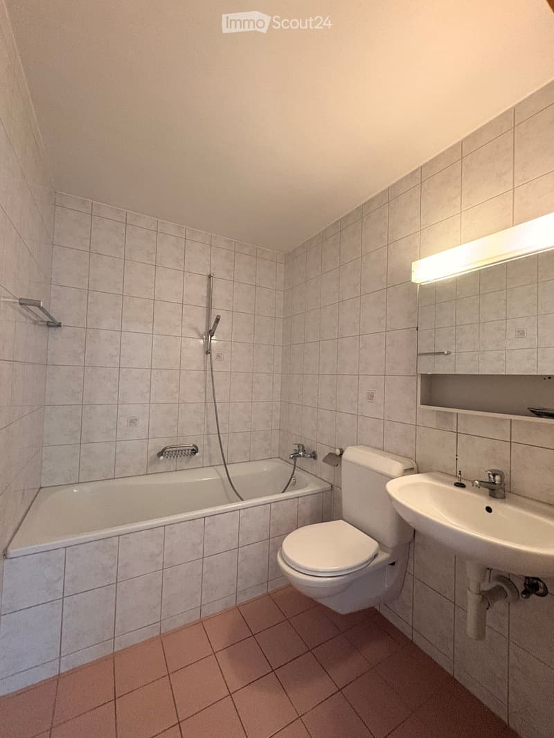 Bathroom with tiled walls and floor, featuring a bathtub, toilet, sink, and mirror with built-in storage.