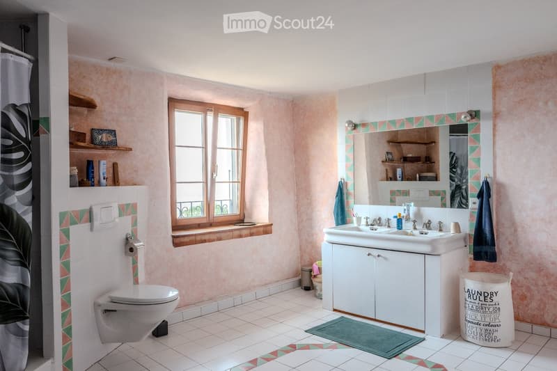 Bright bathroom with pink walls, white tiles, and a large window. Features a toilet, sink with a mirror, cabinet, shelves, and a shower curtain.