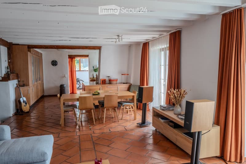 Open-plan living and dining area with wooden furniture, tiled floor, exposed wooden beams, and large windows with curtains.