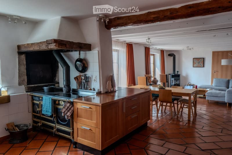 Open-plan kitchen and living area with exposed wooden beams, a large stone fireplace, a wooden kitchen island, a stove, a sink, a wooden dining table with chairs, a sofa, and an ottoman. The space features terracotta tiled flooring and large windows with