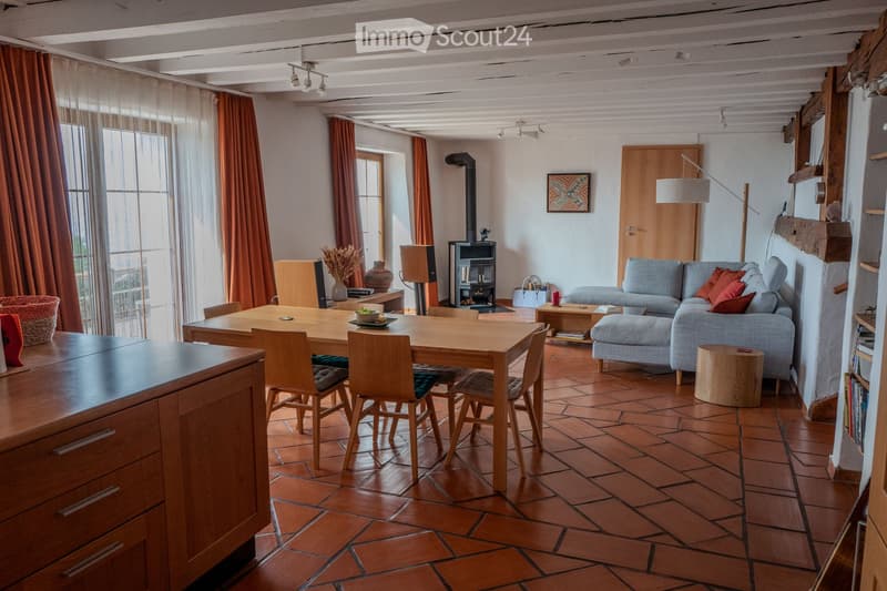 A spacious living room with exposed wooden beams, terracotta tiled floor, and large windows with orange curtains. Features a grey sofa with red cushions, a wooden dining table with chairs, a fireplace, and a floor lamp.