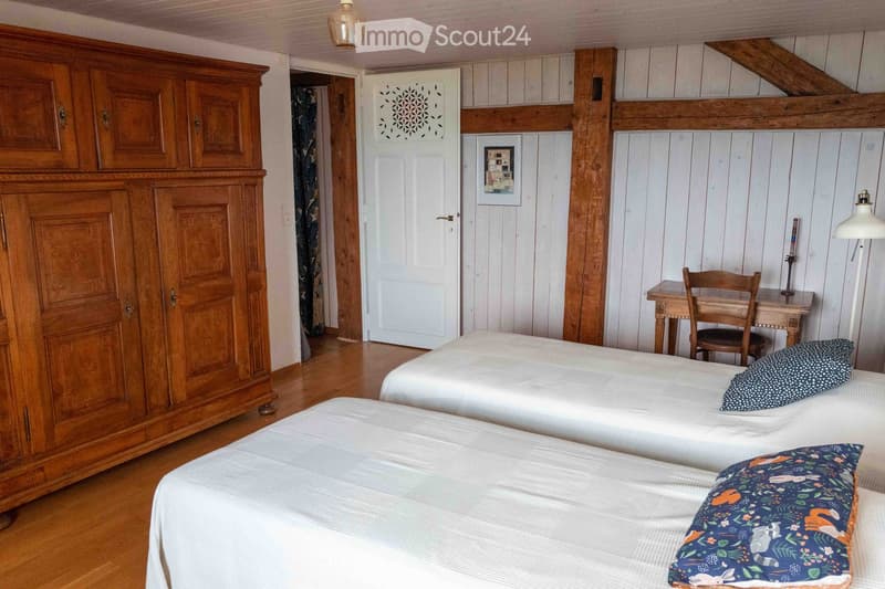 Cozy bedroom with two single beds, a large wooden wardrobe, a small wooden desk with a chair, and a decorative lamp on the wall.