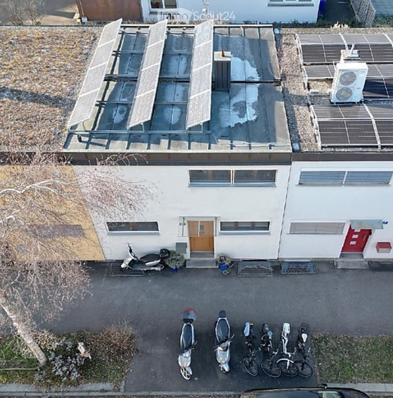 A modern two-story building with a flat roof, white exterior, and multiple windows. The roof features a large solar panel installation. Several scooters and bicycles are parked in front of the building.