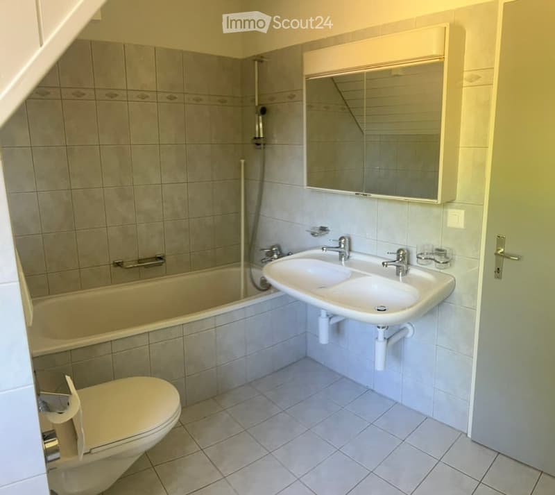 Modern bathroom with a bathtub, toilet, double sink, and a large mirror above the sink. The walls are tiled in a neutral color scheme, and the floor is covered with light-colored tiles.